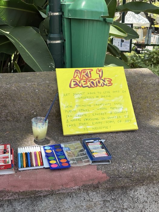 A yellow hand-painted canvas explaining Art 4 Everyone, propped on a stone wall beside watercolours, pastels and pencils.